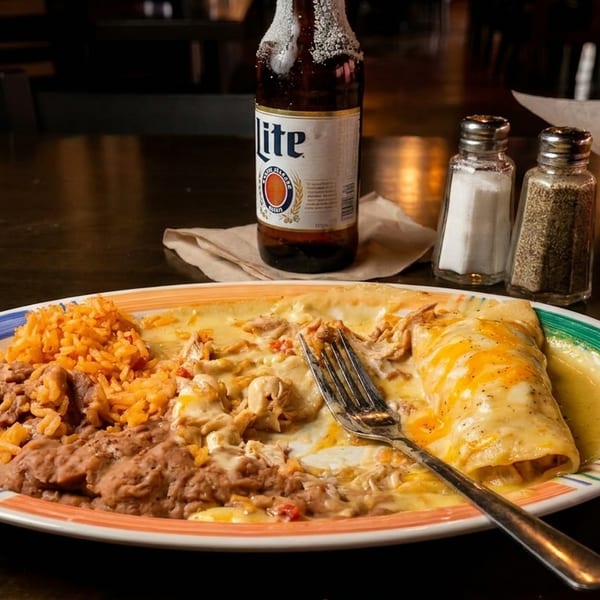 A plate of enchiladas, rice, and beans with a beer and tortilla chips on a dark table.